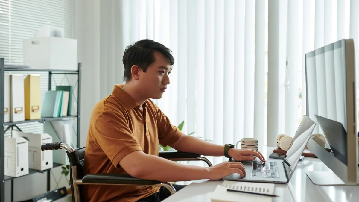 Sales rep in a wheelchair working at a laptop and monitor in a bright office